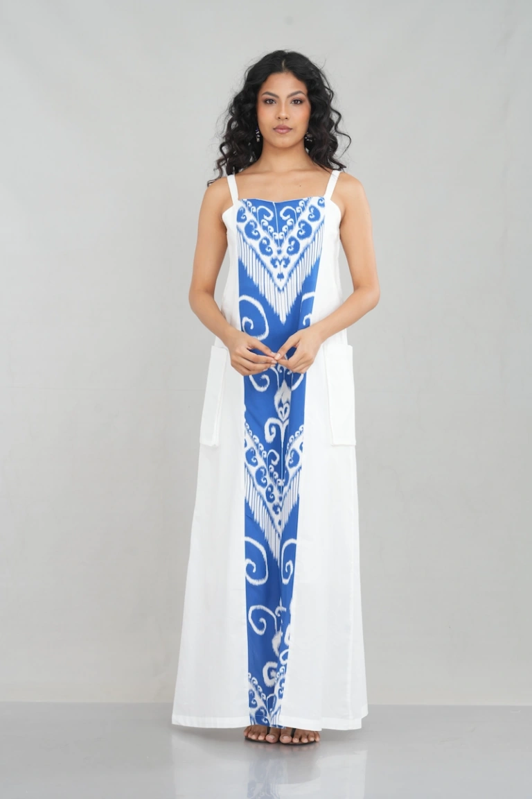 A woman wearing a strappy white maxi dress featuring intricate royal blue embroidery in the front panel, made from a cotton blend fabric, with functional side pockets and a relaxed fit.