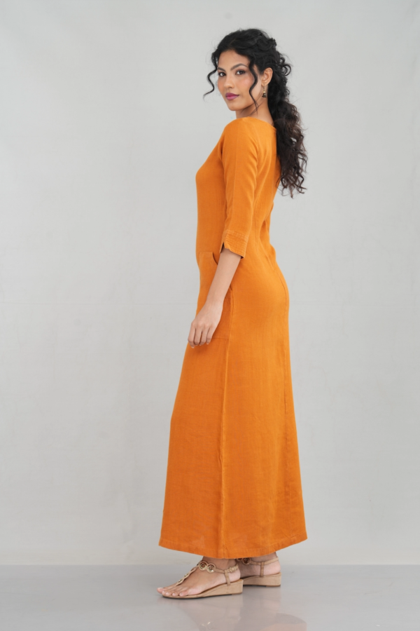 A chic woman wearing a rustic orange linen long sleeve maxi dress with a boat neckline, front pockets, and a tailored fit, styled elegantly with gold-toned sandals.