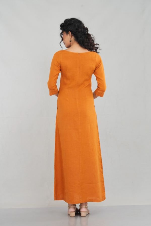 A chic woman wearing a rustic orange linen long sleeve maxi dress with a boat neckline, front pockets, and a tailored fit, styled elegantly with gold-toned sandals.