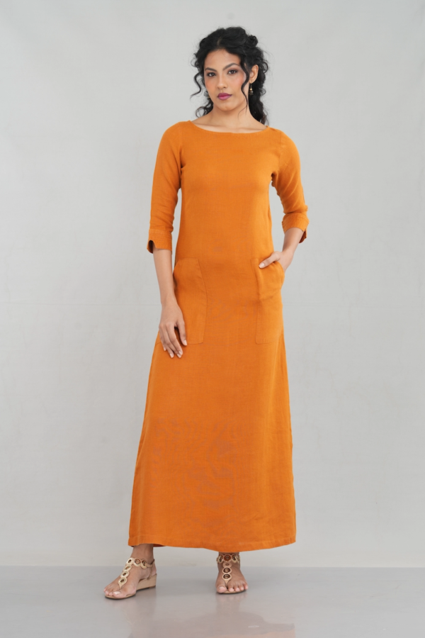 A chic woman wearing a rustic orange linen long sleeve maxi dress with a boat neckline, front pockets, and a tailored fit, styled elegantly with gold-toned sandals.