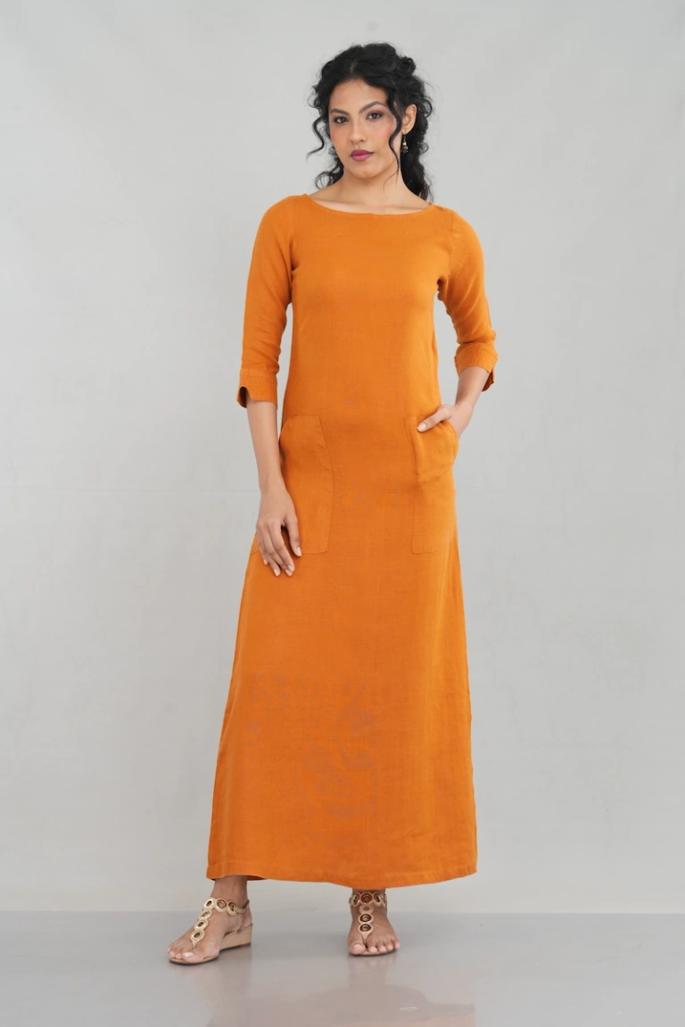 A chic woman wearing a rustic orange linen long sleeve maxi dress with a boat neckline, front pockets, and a tailored fit, styled elegantly with gold-toned sandals.