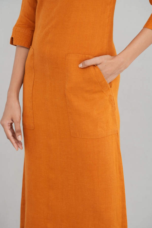 A chic woman wearing a rustic orange linen long sleeve maxi dress with a boat neckline, front pockets, and a tailored fit, styled elegantly with gold-toned sandals.