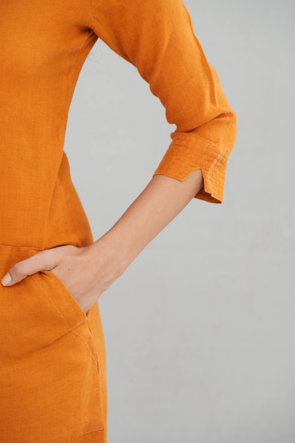 A chic woman wearing a rustic orange linen long sleeve maxi dress with a boat neckline, front pockets, and a tailored fit, styled elegantly with gold-toned sandals.