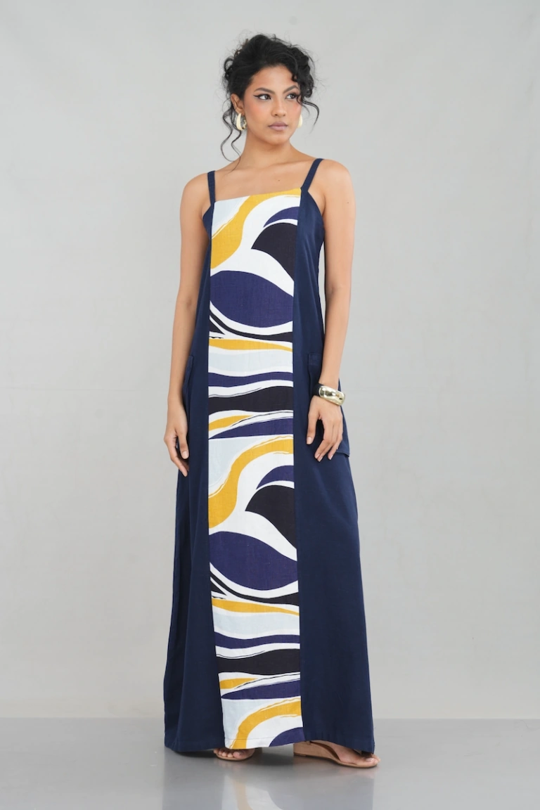 A woman wearing a sleeveless navy blue maxi dress featuring a bold multicolor abstract print in the front panel, crafted from a linen-cotton blend, with a relaxed fit and functional pockets.