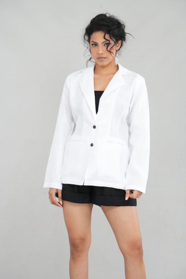 A chic woman wearing a pure white oversized blazer made of linen, featuring a tailored lapel, two-button closure, and functional front pockets, styled elegantly with black shorts and heels.