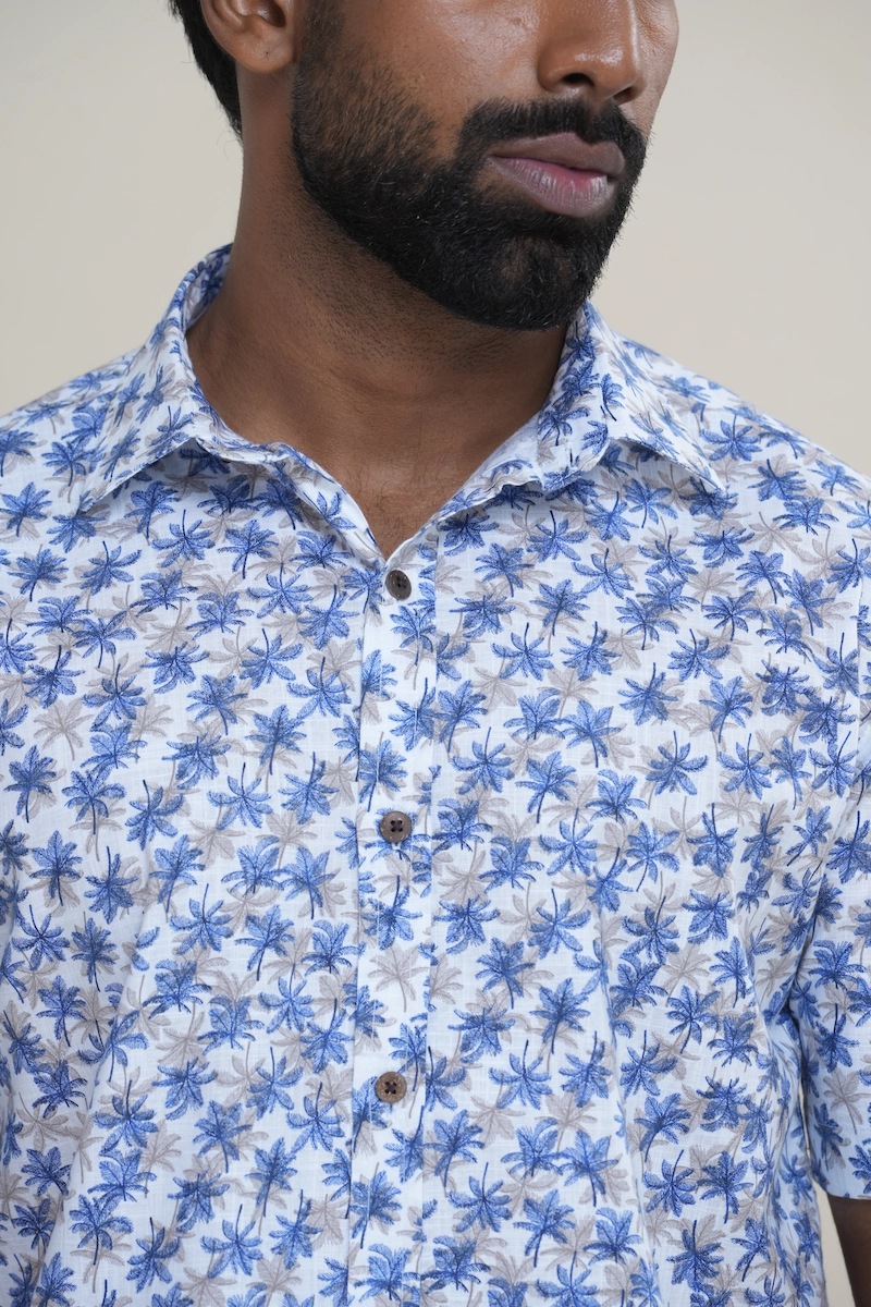 Man wearing a white cotton short sleeve shirt with a white and blue palm tree print, classic collar, and coconut buttons