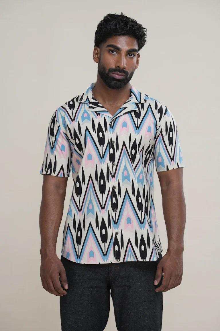 Close-up of a men's linen cotton blend Cuban collar short sleeve shirt with a cream base and a black, blue, and pink abstract geometric print, showing coconut buttons.