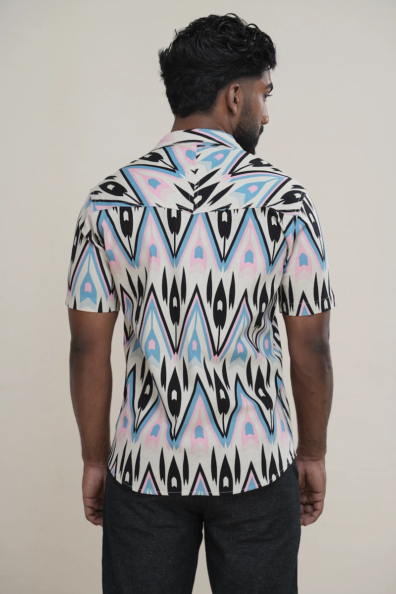 Close-up of a men's linen cotton blend Cuban collar short sleeve shirt with a cream base and a black, blue, and pink abstract geometric print, showing coconut buttons.