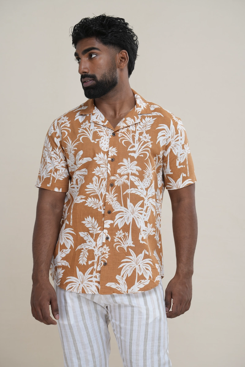 Man wearing a terracotta linen cotton blend Cuban collar shirt with a white pineapple and palm tree print. Close-up of a terracotta and white tropical print Cuban collar shirt highlighting the print detail and collar style.