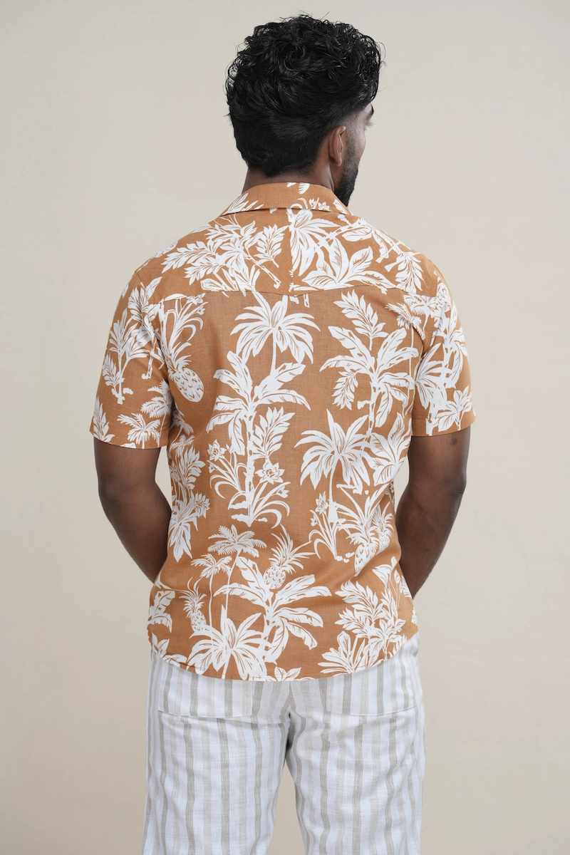 Man wearing a terracotta linen cotton blend Cuban collar shirt with a white pineapple and palm tree print. Close-up of a terracotta and white tropical print Cuban collar shirt highlighting the print detail and collar style.