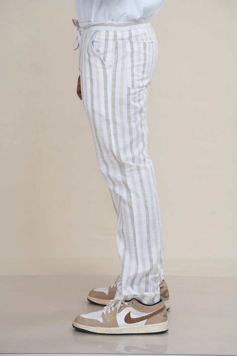 Man wearing off-white and beige striped linen drawstring trousers with a relaxed fit.Close-up of the waist of striped linen trousers, showing the drawstring tie and button closure detail.