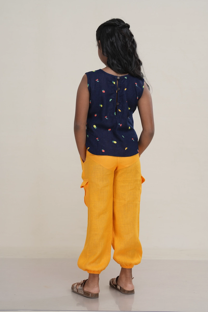 Young girl wearing a navy blue linen-cotton blend sleeveless top with a multi-color fruit print and a full button-back closure. girl wearing a fruit print sleeveless top, showing the full button-down detail.