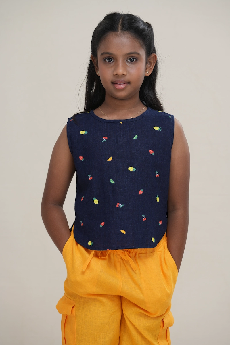 Young girl wearing a navy blue linen-cotton blend sleeveless top with a multi-color fruit print and a full button-back closure. girl wearing a fruit print sleeveless top, showing the full button-down detail.