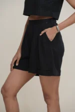 Woman wearing classic black linen pleated high-waist shorts with a cuffed hem and side pockets, the pleat and pocket detail on black high-waisted linen shorts.