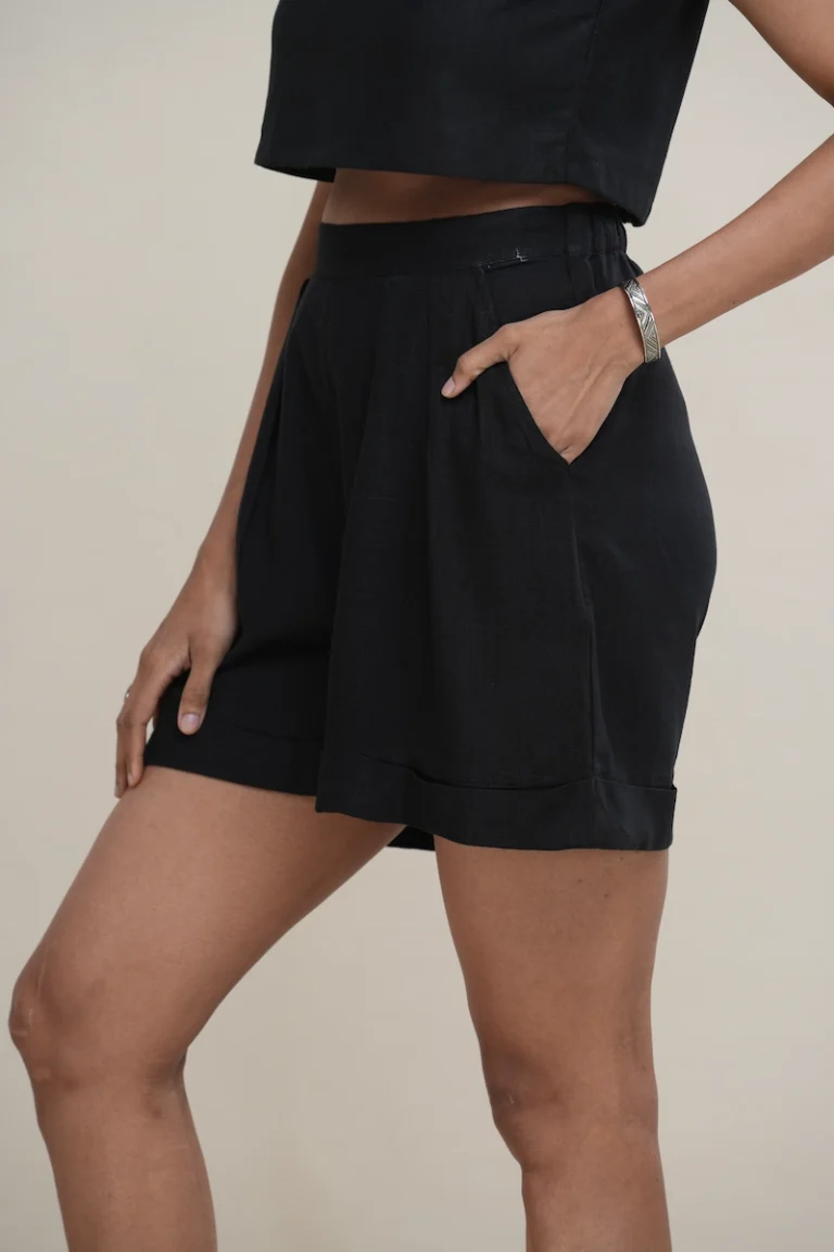 Woman wearing classic black linen pleated high-waist shorts with a cuffed hem and side pockets, the pleat and pocket detail on black high-waisted linen shorts.