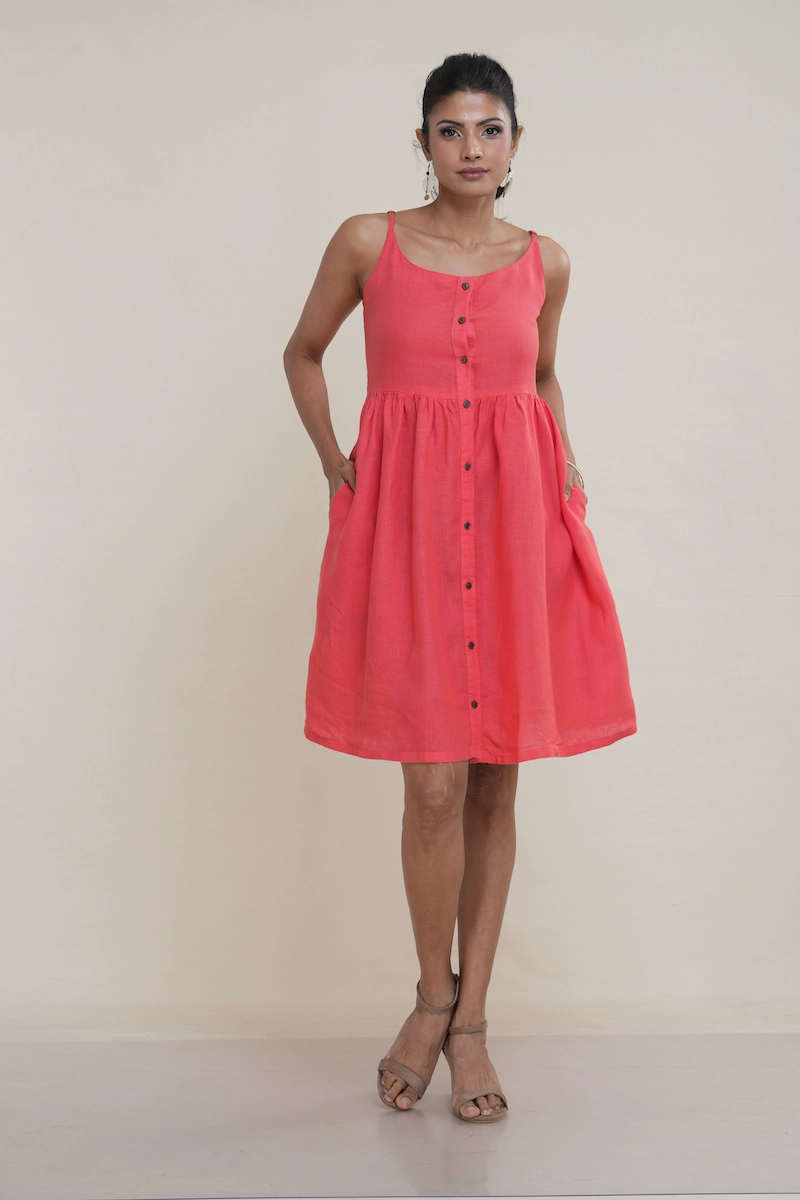 Woman wearing a knee-length coral pink linen button-front cami dress with adjustable straps and side pockets.