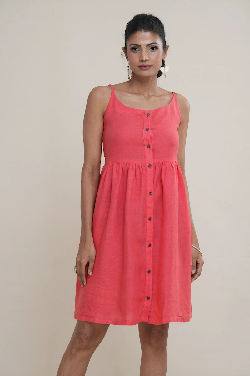 Woman wearing a knee-length coral pink linen button-front cami dress with adjustable straps and side pockets.