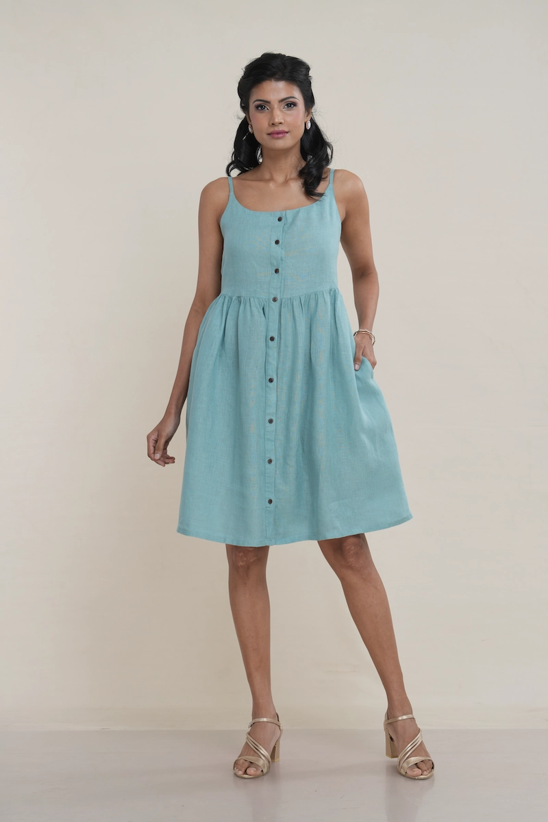 Woman wearing a knee-length seafoam green linen button-front cami dress with side pockets. green linen dress showing adjustable straps and elasticated back panels for a comfortable fit.