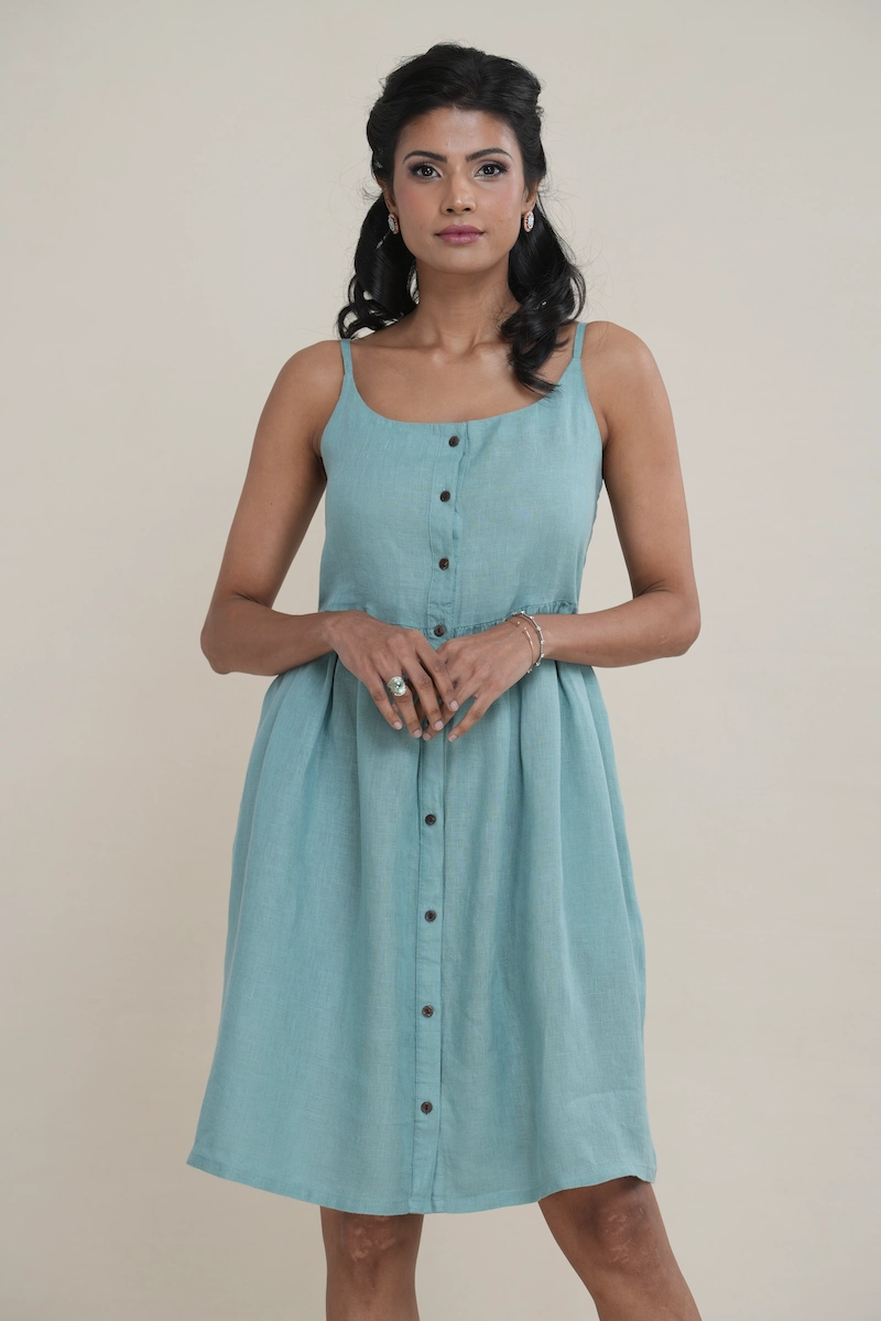 Woman wearing a knee-length seafoam green linen button-front cami dress with side pockets. green linen dress showing adjustable straps and elasticated back panels for a comfortable fit.