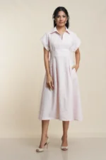 Front view of pale peach linen midi dress with collar, short sleeves, pleated skirt and pockets on model against beige backdrop