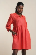 Women's empire waist dress in soft coral red linen with V-neck, long sleeves, and gathered skirt, modeled on beige background